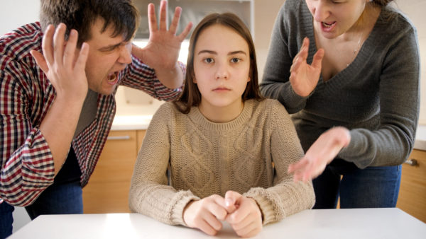 Father and mother shouting and screaming at teenage daughter sitting behind table on kitchen. Family violence, conflicts and relationship problems.